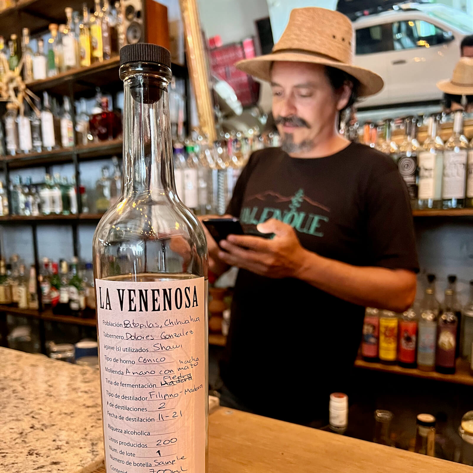 A rare bottle of La Venenosa Raicilla with Esteban Morales out of focus in the background against a large bar